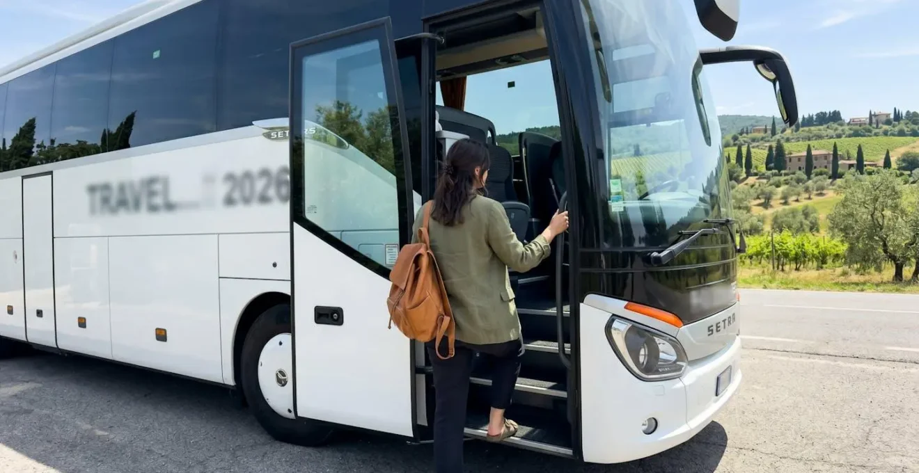 A traveller in casual clothing steps onto a contemporary tour bus, viewed from the side, with sunlit Tuscan hills visible in the background